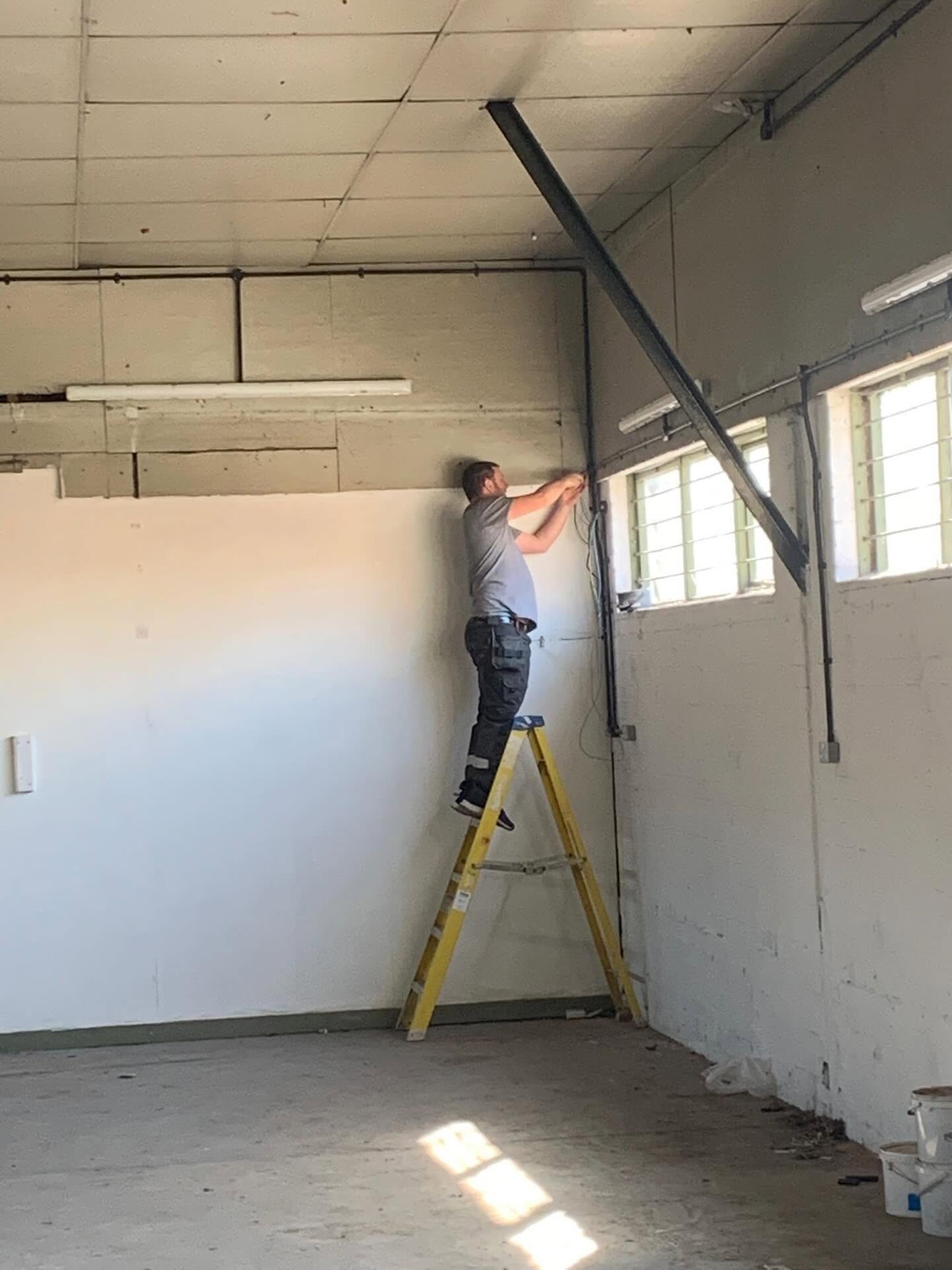 Electrician on a ladder working in a stripped-out warehouse unit